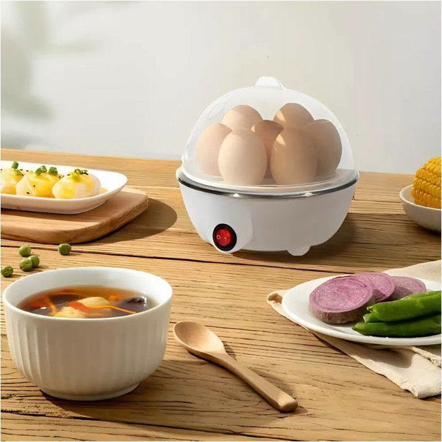 A white electric egg cooker with a clear lid sits on a wooden table, steaming several eggs. Surrounding it are bowls of soup, sliced purple sweet potato, green beans, and corn on a plate, with a wooden spoon in the foreground.