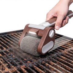A hand holds a brown and silver grill cleaning roller brush with an integrated light, scrubbing a dirty grill grate over a white background.